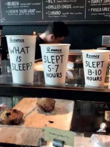 Fotografia de três copos de café de tamanhos diferentes dispostos em balcão de cafeteria. No copo grande (16 ounce) está escrito 'WHAT IS SLEEP?'. No copo médio (12 ounce) está escrito 'SLEPT 5-7 HOURS'. No copo pequeno (8 ounce) está escrito 'SLEPT 8-10 HOURS'
