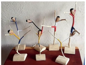Fotografia de esculturas de bailarinos entalhados em gravetos finos de madeira, com corpos alongados em poses de dança, fixados sobre pequenas bases de madeira clara