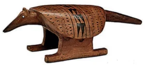 Fotografia de um banco indígena de madeira esculpido na forma de um tatu, com cabeça alongada, cauda e casco decorado com grafismos geométricos pirogravados.