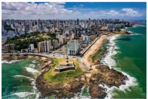 Fotografia aérea da orla de Salvador (BA), com o Farol da Barra em primeiro plano sobre uma ponta verde junto ao mar e, ao fundo, grande concentração de edifícios altos e verticalizados ao longo da costa.