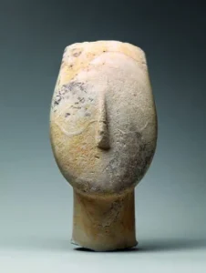 Fotografia de escultura anônima em mármore representando uma cabeça feminina estilizada, alongada e oval, de superfície lisa e clara, com nariz proeminente em relevo e ausência de olhos e boca. Circa 2700-2500 a.C., 8 × 3,2 cm, Metropolitan Museum of Art, Nova Iorque