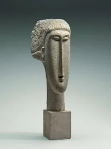 Fotografia da escultura 'Cabeça de mulher', de Modigliani, em calcário. Rosto alongado e geometrizado, com nariz reto e comprido, olhos e boca sugeridos por incisões e cabelo indicado por sulcos verticais, apoiada sobre base cúbica. Circa 1910-1911, 68,3 × 15,9 × 24,1 cm, National Gallery of Art, Washington